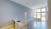 This contemporary single-floor space offers a clean, modern aesthetic defined by smooth polished concrete floors and cool-toned blue-gray walls that lend an airy, minimalist feel throughout. The open layout is anchored by a sleek kitchenette island finished with speckled granite countertops, a stainless steel sink, and integrated cabinetry, providing both functionality and a refined focal point. Directional track lighting and wall-mounted accent lights are thoughtfully positioned to illuminate the space evenly, complementing the abundant natural light that pours in through large, floor-to-ceiling windows along one wall. These expansive windows not only brighten the interior but also offer views of the surrounding urban environment, creating a strong visual connection between the indoors and outdoors. The high ceilings enhance the sense of openness and volume, contributing to the overall spacious and uncluttered atmosphere. The restrained color palette and durable finishes throughout suggest a space well-suited for a variety of uses, from a professional studio or creative workspace to a modern live-work environment. Every element of the design reflects a contemporary, functional approach with an emphasis on clean lines and quality materials.
