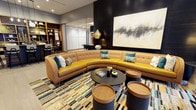 This single-floor lounge space showcases a thoughtfully curated contemporary interior that balances bold aesthetics with comfortable functionality. The focal point of the room is a sweeping curved sectional sofa upholstered in warm mustard-toned leather, accented with teal and neutral decorative pillows that introduce a vibrant color palette throughout the space.

A multicolored area rug in tones of blue, gold, and cream anchors the seating area, complementing the overall design scheme. Cylindrical side tables with wooden tops and dark bases are arranged around a sleek circular black coffee table, offering both style and practicality.

A large-scale abstract artwork with dynamic brushwork in black, white, and gold commands attention on the dark feature wall, adding an artistic dimension to the space. A brushed gold pendant light hangs above, contributing warmth and a touch of glamour.

Recessed lighting illuminates the space evenly, while an open shelving unit with warm wood tones is visible in the background, alongside a semi-private area with bar-height seating, suggesting a multifunctional layout. The overall ambiance is sophisticated, modern, and inviting.
