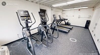Located on Carolina Ave in Durham, NC, this fitness center is organized across two connected exercise rooms, offering a well-rounded workout environment. The space features durable rubber flooring throughout, providing both safety and functionality for a variety of exercise activities. Fluorescent and panel lighting illuminates the exposed structural ceiling, giving the facility a practical, no-frills aesthetic.

The primary cardio room is equipped with multiple treadmills and elliptical machines, providing ample options for cardiovascular training. A sliding glass door in this room allows access to an exterior area, introducing natural light into the space. An adjacent room houses a Matrix functional trainer with cable attachments and a full rack of dumbbells arranged on a three-tier stand, catering to strength and resistance training needs. Stability balls in multiple sizes are also available, supported on a dedicated rack alongside exercise mats, supporting flexibility and core training routines.

A pass-through doorway connects the two rooms seamlessly, allowing users to transition between cardio and strength training with ease. A wall clock ensures users can manage their workout time effectively. The overall design is utilitarian and efficient, prioritizing function and equipment capacity within a compact footprint.