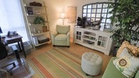 This two-floor space exudes a warm, cottage-inspired charm that invites comfort and relaxation. The interior design leans into a soft, nature-inspired color palette, with furnishings and décor carefully chosen to create an inviting and cohesive aesthetic throughout.

The main living area showcases a floral-upholstered accent chair paired with a tufted round ottoman, both resting atop a richly striped area rug in earthy tones of green, coral, and tan. A lean-to ladder shelf occupies one corner, displaying decorative jars, a potted plant, and woven baskets, adding a rustic, layered feel to the space. A classic table lamp provides warm ambient lighting, complementing the room's inviting glow.

A white cottage-style media console serves as a functional centerpiece, outfitted with glass-panel cabinet doors and open shelving that display colorful bowls and decorative items. A large multi-pane mirror rests above it, enhancing the sense of depth and light in the room.

The overall ambiance is relaxed and thoughtfully curated, blending functional pieces with decorative touches to create a space that feels both lived-in and carefully styled. The two-floor layout provides additional space to explore, promising a consistent attention to detail and comfort throughout the entire property.