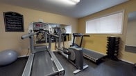 Located at 621 Westover Hills Blvd in Richmond, Virginia, this single-story amenity facility offers two distinct functional areas designed to serve the needs of its users. The layout connects a community lounge space with a dedicated fitness center, providing a well-rounded on-site experience.

The lounge area features warm wood-look flooring and soft gray walls, creating a clean, welcoming atmosphere. Two round dark-wood tables, each surrounded by modern black chairs, offer seating for small gatherings or casual meetings. Framed certificates and artwork line the walls, adding a polished, professional touch. A glass-paned door along one wall invites natural light into the space, enhancing its open and airy feel.

The fitness center is outfitted with rubber flooring for safety and durability. Equipment includes multiple treadmills, an elliptical trainer, a stationary bike, a dumbbell rack with free weights, a weight bench, and a stability ball. Full-length wall mirrors expand the perceived space and allow users to monitor their form during workouts. Windows fitted with blinds provide natural light while maintaining privacy. A posted fitness center rules sign near the entrance reflects an organized, community-minded environment. Together, both areas form a functional and well-maintained shared facility.