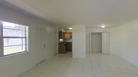 This single-floor space offers a straightforward, open layout that connects the main living area to the kitchen and an additional room. The living area is generously sized, finished with light-toned ceramic tile flooring that extends throughout, contributing to a clean and cohesive look. White walls and flush-mount ceiling fixtures provide a bright, neutral backdrop throughout the space.

The kitchen is positioned off the main living area and features warm-toned wooden cabinetry on both the upper and lower levels, dark countertops, and a built-in appliance. A small window above the counter allows in natural light. The kitchen's layout is compact yet functional, with adequate storage provided by the full set of upper and lower cabinets.

Adjacent to the main living space, a secondary room is accessible through a wide doorway fitted with bi-fold doors, offering flexibility in its use. The space benefits from a simple, practical design with consistent finishes across all areas. Two entry doors are visible in the main living area, providing multiple points of access. Overall, the space presents a clean, utilitarian character suited for everyday living.
