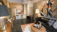 This single-floor space showcases a thoughtfully designed open-concept layout that integrates the living, dining, and kitchen areas into one cohesive environment. The interior features elevated ceilings that enhance the sense of spaciousness throughout. Dark polished concrete flooring grounds the space, providing a sleek, industrial contrast to the warm, earthy tones of the walls and furnishings.

The living area is anchored by a tufted leather ottoman coffee table and a bold black leather armchair, complemented by an orange Moroccan-patterned area rug with a plush sheepskin overlay — adding texture and visual interest. A large abstract artwork commands one wall, contributing to the space's eclectic character. A sculptural metallic wall installation serves as a striking decorative accent between the living and dining zones.

The compact dining area accommodates intimate gatherings with a small table and modern chairs. The kitchen features a peninsula with bar-height seating, stainless steel appliances including a full-size refrigerator, warm wood cabinetry, and a pendant light, creating a functional yet stylish culinary space. Overall, the unit exudes a contemporary, design-forward ambiance with a rich, layered aesthetic.