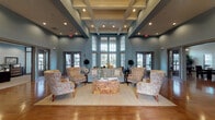 This single-story community space makes a strong architectural impression through its dramatically tall ceilings, coffered detailing, and an abundance of natural light streaming through large, multi-pane windows that rise nearly to the roofline. The warm, blue-gray walls provide a calm, cohesive backdrop throughout the interior, complemented by rich hardwood flooring that extends across all areas.

The central lounge area serves as the heart of the space, featuring a carefully arranged seating arrangement of floral-patterned armchairs and a sofa surrounding a distinctive wood-inlay coffee table, all anchored on a neutral area rug. This inviting arrangement encourages conversation and relaxation in equal measure.

Adjacent seating nooks near the entry points offer additional intimate areas, furnished with wicker and upholstered chairs alongside decorative topiary plants and table lamps that add warmth. Glazed double-door entryways on multiple sides provide clear views to the outdoors while maximizing natural light penetration.

A separate room, visible through sliding glass-panel doors, appears to function as a private meeting or office area, outfitted with a chandelier and desk furniture. Recessed lighting throughout supplements the natural illumination, ensuring the entire space remains bright and welcoming at all times.