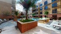 This expansive outdoor courtyard is designed as a resort-style amenity space nestled within a multi-story residential complex. The architecture blends warm brick masonry at the lower levels with smooth stucco facades on the upper floors, creating a visually inviting enclosure that frames the entire outdoor area.

At the heart of the courtyard lies a sparkling swimming pool complemented by an elegant fountain feature, adding both visual interest and a soothing ambiance. Raised brick planters filled with lush tropical greenery, including mature palm trees, serve as natural focal points throughout the space. Generous stone-tiled walkways connect the various zones, providing easy circulation across the courtyard.

A well-appointed sun deck lined with cushioned lounge chairs and patio umbrellas offers ample space for relaxation, while shaded seating areas with tables and chairs accommodate outdoor dining and socializing. Thoughtfully designed garden beds and ornamental plantings line the building perimeters, featuring a mix of tropical shrubs, ornamental grasses, and seasonal flowers that add vibrant color and texture.

A dedicated recreational lawn area provides space for outdoor games, with a cornhole set visible on the neatly manicured grass. Low-profile bollard lighting is integrated throughout the pathways, ensuring the courtyard remains welcoming into the evening hours. Overall, the space exudes a lively yet relaxed open-air atmosphere perfect for outdoor living.