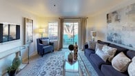 This single-floor residence offers a thoughtfully designed layout encompassing a living room, dining area, kitchen, two bedrooms, and multiple bathrooms. The living room serves as the central gathering space, featuring carpet flooring, a sliding glass door that opens to a private outdoor patio, and a adjacent window that brings in generous natural light. A comfortable seating arrangement and glass coffee table anchor the room, while a wall-mounted television and console table complete the space.

The dining area flows naturally from the living room, featuring carpet flooring and a decorative round mirror accent. The kitchen is equipped with light wood-toned cabinetry, ample counter space, a double sink, and white appliances including a range, microwave, dishwasher, and refrigerator. Wood-look flooring adds warmth to this functional space.

Both bedrooms are carpeted and include ceiling fans. The primary bedroom features a rustic wood-panel headboard and a large dresser with mirror, while the secondary bedroom includes a built-in desk area and closet. The bathrooms feature wood-look flooring, one with a tub/shower combination and another with a glass-enclosed shower stall and vanity.