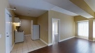 This single-floor space features a straightforward, functional layout with a clear visual connection between the main living area and the kitchen. The walls are finished in a warm golden-beige tone, creating a consistent and cohesive look throughout the space.

The living area is laid with rich dark hardwood flooring, which transitions into light ceramic tile in the kitchen zone, clearly delineating the two areas. The kitchen is equipped with a white refrigerator and a stove, with the tile flooring providing a practical and easy-to-maintain surface. A doorway with white trim leads from the main space into an adjacent room that benefits from natural light, suggesting a window or exterior-facing feature within.

White baseboards and door trim contrast cleanly against the warm wall color, adding a crisp, finished appearance. The ceiling features a textured finish and includes visible structural beams or soffits, adding subtle architectural definition to the space. The overall ambiance is modest and utilitarian, with a neutral palette that offers a blank canvas for personalization.