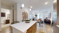 This contemporary single-floor residence showcases a thoughtfully designed open-concept layout that seamlessly connects the kitchen, dining, and living areas. The kitchen is a standout feature, equipped with warm wood-toned cabinetry, stainless steel appliances including a range, microwave, and French door refrigerator, complemented by crisp white quartz countertops. A generously sized center island with seating provides both additional prep space and a casual dining option, illuminated by modern pendant lighting overhead. Track lighting and recessed fixtures throughout ensure well-lit interiors. Light-toned hardwood flooring flows throughout the main living areas, enhancing the bright and airy atmosphere. The living room offers an inviting setting with a ceiling fan, large windows, and glass doors that invite natural light and outdoor views. The bedroom features plush carpeting, ample window coverage with blinds, and comfortable proportions. The bathroom is elegantly finished with large-format tile flooring, a floating vanity with ample storage, a wide illuminated mirror, and a glass-enclosed walk-in shower. Clean white walls and modern fixtures throughout the space contribute to a cohesive, contemporary aesthetic that balances style and functionality.