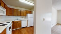 This single-floor residence offers a practical and comfortable layout across several well-defined living areas. The kitchen is a standout feature, outfitted with warm oak cabinetry that provides generous upper and lower storage, white appliances including a coil electric range, over-the-range microwave, dishwasher, and refrigerator, as well as light-toned countertops and hardwood flooring that adds a touch of warmth. The kitchen opens toward the main living areas, creating a connected feel throughout the home. The living room is a bright, open space with plush wall-to-wall carpeting, a ceiling fan, textured ceilings, and a large sliding glass door that invites natural light and provides views of the surrounding greenery. A baseboard heating system runs along the perimeter. The bedroom features the same soft carpeting, a sliding-door closet for convenient storage, and a baseboard heater. The bathroom includes a wood-toned vanity with a single sink, light countertop surface, a large mirror, tile flooring, and an open closet area offering additional storage space. Overall, the space presents a clean, functional interior with consistent finishes throughout.