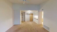 This single-floor unit offers a well-organized layout across one bedroom and one bathroom. The main living area is finished with wall-to-wall neutral carpeting and painted in a soft, light blue-gray tone that lends a calm, airy atmosphere throughout. A ceiling fan is centrally mounted overhead, providing both airflow and a subtle focal point to the room.

The open-concept design connects the living area to the dining space through a wide arched passageway, enhancing the sense of flow and openness. A kitchen pass-through bar with a pendant light above serves as both a functional surface and a visual divider between the kitchen and the adjacent dining or living area, allowing light and sightlines to carry through the space.

Toward the rear of the living area, a large sliding wood-panel door provides access to a closet with built-in shelving, offering generous storage. The entryway is positioned conveniently off to one side, with a coat closet nearby. The bathroom, visible through an adjacent doorway, features warm wood-toned cabinetry. The overall layout is practical and efficient, making good use of the available square footage.