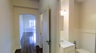 This single-floor space showcases timeless architectural character throughout its layout. The hallway serves as the central corridor, finished with deeply stained, highly polished hardwood floors that reflect natural light and add warmth and elegance to the interior. A graceful arched doorway frames the passage leading toward a bright, naturally lit room at the end of the hall, where large windows allow abundant daylight to pour in. A classic radiator beneath the windows hints at traditional heating infrastructure consistent with older construction. The walls are finished in a clean, neutral cream tone that complements the dark flooring and white trim work. Doors along the hallway feature classic hardware, reinforcing the period character of the space. Adjacent to the hallway, a bathroom is partially visible, appointed with a white vanity sink, a wall-mounted mirror, and a sconce light fixture with frosted glass shades that provide warm, functional lighting. The overall ambiance of the space combines historic architectural charm with clean, well-maintained finishes, creating an inviting and functional interior well-suited for comfortable everyday living.