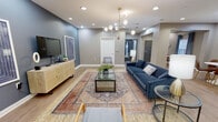 This thoughtfully designed single-floor communal space blends modern aesthetics with functional comfort across several distinct areas. The main lounge features a bold navy blue sofa, layered area rugs, and a striking brass multi-globe chandelier, all set against cool grey-blue accent walls and light hardwood-style flooring. Recessed lighting and a wall-mounted television anchor the space, complemented by a rattan media console that adds a warm, organic touch.

Adjacent to the main lounge, a secondary sitting area offers a more intimate setting with a grey tufted sofa, a geometric brass pendant light, and natural light streaming through windows dressed in soft blue curtains. A mid-century-inspired round white dining table with cognac leather chairs occupies a bright dining nook, illuminated by a sculptural gold pendant fixture and a decorative sunburst wall accent.

The fully equipped kitchen is well-appointed with warm honey-toned cabinetry, granite countertops, a tile backsplash, stainless steel appliances including a refrigerator and microwave, a coffee and beverage station, and a central island. The open, column-accented layout connects each zone seamlessly, creating a cohesive and inviting environment suited for relaxation, socializing, or casual gatherings.