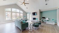 Located at 70 Greenway Square in Dover, Delaware, this single-story community clubhouse is a refined and welcoming gathering space defined by elegant architectural details and a cohesive coastal-inspired color palette. The entry foyer makes an immediate impression with its arched ceiling, decorative chandelier, and stately white columns framing the passage into the main lounge area. Underfoot, richly veined hardwood-style flooring flows throughout, complementing the soft gray and teal tones that define the interior. The main lounge is anchored by a dramatic vaulted ceiling with exposed ridgeline and a ceiling fan, while a wall of arched, grid-pane windows floods the room with natural light and offers views of the surrounding community. A white-painted stone fireplace serves as a focal point, topped with a mounted flat-screen television, with a dedicated workstation neatly positioned nearby. Plush teal seating arrangements invite residents to relax or gather socially. A secondary sitting area with a glass-top table and teal accent chairs provides a quieter retreat beneath its own vaulted window wall. The space also includes a functional kitchen featuring white cabinetry, stainless steel appliances, a breakfast bar with seating, and teal accent walls that tie seamlessly into the overall design aesthetic.