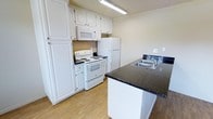 This single-floor space centers around a well-organized kitchen that combines practicality with clean aesthetics. The kitchen is outfitted with white raised-panel cabinetry that extends to the upper walls, providing generous storage space. Dark granite countertops offer a contrasting visual element against the white cabinetry and appliances, adding a touch of elegance to the room.

The kitchen is equipped with a full suite of white appliances, including an electric coil range, an over-the-range microwave, and a full-sized refrigerator. A peninsula with matching dark granite extends into the space, incorporating a double-basin stainless steel sink with a standard faucet, and creating a natural division within the kitchen area.

Overhead fluorescent lighting provides bright, even illumination throughout the kitchen. The flooring consists of light-toned wood-look material, contributing to a warm and cohesive atmosphere. The neutral wall tones complement the overall color palette, resulting in a clean, functional, and inviting kitchen environment.
