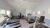 This dedicated fitness center offers a comprehensive workout environment designed to support a wide range of exercise routines. The space is defined by its vaulted ceilings, which lend an open, airy feel throughout. Walls are painted in a calming two-tone blue and white scheme, with a clean white chair rail dividing the upper and lower portions, creating a tidy and refreshing atmosphere.

Natural light pours in through multiple large windows along one wall, offering pleasant views of the surrounding greenery and outdoor recreational areas. A set of glass double doors provides direct access to the outdoors, further enhancing the connection to the outside environment. Ceiling fans and recessed lighting ensure comfortable conditions regardless of the time of day.

The fitness floor is carpeted and well-organized, accommodating multiple treadmills, elliptical trainers, a stationary bike, and a multi-station strength training machine. A large wall mirror on one side expands the sense of space while allowing users to monitor their form. A wall-mounted television is conveniently positioned for entertainment during workouts. Safety features, including a fire extinguisher and emergency exit signage, are also present. The overall layout is practical, spacious, and thoughtfully arranged to support a variety of fitness goals.
