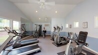 This dedicated fitness center offers a comprehensive workout environment designed to support a wide range of exercise routines. The space is defined by its vaulted ceilings, which lend an open, airy feel throughout. Walls are painted in a calming two-tone blue and white scheme, with a clean white chair rail dividing the upper and lower portions, creating a tidy and refreshing atmosphere.

Natural light pours in through multiple large windows along one wall, offering pleasant views of the surrounding greenery and outdoor recreational areas. A set of glass double doors provides direct access to the outdoors, further enhancing the connection to the outside environment. Ceiling fans and recessed lighting ensure comfortable conditions regardless of the time of day.

The fitness floor is carpeted and well-organized, accommodating multiple treadmills, elliptical trainers, a stationary bike, and a multi-station strength training machine. A large wall mirror on one side expands the sense of space while allowing users to monitor their form. A wall-mounted television is conveniently positioned for entertainment during workouts. Safety features, including a fire extinguisher and emergency exit signage, are also present. The overall layout is practical, spacious, and thoughtfully arranged to support a variety of fitness goals.