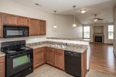 This single-floor residence offers a well-designed open-concept layout that seamlessly connects the main living areas. The living room is a standout space, highlighted by soaring vaulted ceilings that amplify the sense of openness, gleaming hardwood floors, and a tile-surround fireplace framed by built-in shelving. Large windows flood the room with natural light, while a sliding glass door provides direct access to a private balcony with exterior views.

The kitchen is thoughtfully appointed with rich medium-toned cabinetry, dark granite-style countertops, and a full suite of black appliances including an electric range, over-the-range microwave, dishwasher, and refrigerator. A peninsula with a double sink and pendant lighting above serves as a functional divider between the kitchen and living area. Tile flooring grounds the kitchen space.

A carpeted bedroom features a ceiling fan and a sliding glass door offering additional outdoor access and natural light. The bathroom is practical and spacious, finished with tile flooring, a wooden vanity with a white countertop, and notably, a stacked washer and dryer unit conveniently located within the space. A walk-in closet is accessible directly from the bathroom, offering generous storage. The overall ambiance is clean, functional, and comfortable throughout.