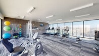Located at 501 Virginia Ave in Indianapolis, Indiana, this fitness center offers a thoughtfully designed exercise space ideal for a comprehensive workout experience. The room is defined by a striking contrast of materials: exposed brick walls in a neutral gray tone anchor one side of the space, while expansive floor-to-ceiling windows along the opposite wall flood the interior with natural light and frame sweeping views of the Indianapolis skyline and surrounding cityscape.

The flooring throughout features a modern striped carpet in varied gray tones, lending a contemporary aesthetic while providing comfort underfoot. Sleek linear LED fixtures run across the white ceiling, ensuring bright, even illumination throughout the day and evening.

The space is fully outfitted with a wide array of fitness equipment, including multiple treadmills, elliptical trainers, and stationary bikes for cardio workouts. A multi-function power tower supports strength and bodyweight training, while a dedicated free-weight area includes a full dumbbell rack, a weight bench, and medicine balls in multiple sizes. A wall-mounted television provides entertainment during workouts.

The open, airy layout allows ample room to move freely between equipment, while full-length mirrors enhance the sense of space. The combination of natural light, city views, modern finishes, and diverse equipment makes this fitness center a functional and inviting environment for all levels of exercise.