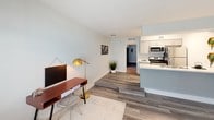 This single-floor unit offers a clean, modern aesthetic throughout, highlighted by durable wood-look flooring in cool gray tones that flows seamlessly across the living spaces. The open-concept layout connects the living area to the kitchen, creating a functional and cohesive floor plan. The living space is bright and welcoming, with natural light entering through a window fitted with horizontal blinds, complemented by neutral, light-toned walls that enhance the airy feel of the room. A subtle step separates the living area from the kitchen zone, adding a touch of architectural interest. The galley-style kitchen is well-appointed with white shaker cabinetry, light quartz-style countertops, and a full suite of stainless steel appliances including a range, over-the-range microwave, refrigerator, and dishwasher. A double-basin stainless steel sink sits beneath a clean accent wall in soft blue, adding a gentle pop of color. A hallway connects the main living areas to the bedroom and bathroom. The bedroom is generously sized, featuring the same wood-look flooring and a window that invites natural light. The bathroom includes a full tub-and-shower combination with built-in shelving, brushed nickel fixtures, and a clean white finish throughout, providing a functional and refreshing space.