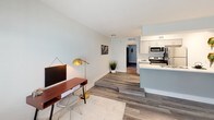 This single-floor unit offers a clean, modern aesthetic throughout, highlighted by durable wood-look flooring in cool gray tones that flows seamlessly across the living spaces. The open-concept layout connects the living area to the kitchen, creating a functional and cohesive floor plan. The living space is bright and welcoming, with natural light entering through a window fitted with horizontal blinds, complemented by neutral, light-toned walls that enhance the airy feel of the room. A subtle step separates the living area from the kitchen zone, adding a touch of architectural interest. The galley-style kitchen is well-appointed with white shaker cabinetry, light quartz-style countertops, and a full suite of stainless steel appliances including a range, over-the-range microwave, refrigerator, and dishwasher. A double-basin stainless steel sink sits beneath a clean accent wall in soft blue, adding a gentle pop of color. A hallway connects the main living areas to the bedroom and bathroom. The bedroom is generously sized, featuring the same wood-look flooring and a window that invites natural light. The bathroom includes a full tub-and-shower combination with built-in shelving, brushed nickel fixtures, and a clean white finish throughout, providing a functional and refreshing space.