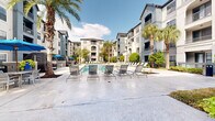 This impressive outdoor amenity space centers around a large, resort-style swimming pool with crystal-clear turquoise water, designed to offer a luxurious retreat within a multi-story residential community. The pool area is generously proportioned, accommodating an extensive arrangement of lounge chairs and sun loungers positioned along its broad concrete decking — ideal for relaxing and sunbathing throughout the day.

Tall, mature palm trees are strategically planted throughout the courtyard, adding a tropical ambiance and providing natural shade across the deck. The surrounding landscape features well-maintained greenery, neatly trimmed shrubs, and decorative planting beds that enhance the overall aesthetic.

Several distinct seating zones are distributed around the pool, including umbrella-shaded dining tables with chairs and enclosed wicker pod seating, offering a variety of social and private gathering options. Fountain features integrated into the pool add a dynamic water element to the setting.

The courtyard is enclosed by the multi-story residential buildings on all sides, creating a private, inner-courtyard feel while still allowing abundant natural light and open sky views. The architecture of the surrounding buildings features arched windows, balconies, and a neutral stucco exterior, contributing to an upscale, cohesive design. The overall ambiance is bright, open, and resort-like, making this outdoor space a standout communal amenity.
