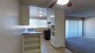 This single-floor space offers a practical and functional layout that seamlessly connects the kitchen and living areas. The kitchen is well-appointed with white cabinetry that extends to the ceiling, providing generous storage space. A dark countertop runs along the kitchen perimeter, complemented by a stainless steel sink positioned beneath a window fitted with vertical blinds that invite natural light into the space. A black gas range with an overhead exhaust hood serves as the cooking centerpiece. An open shelving unit divides the kitchen from the adjacent living area, adding both display and storage functionality while maintaining an open, connected feel. The living area features plush carpeting and continues the natural light theme with additional vertical blinds covering a large window. A ceiling fan with an integrated light fixture is centrally positioned to provide both ventilation and ambient lighting. The textured ceiling and warm neutral wall tones throughout create a cohesive and understated aesthetic. The light-toned flooring in the kitchen provides a clean contrast to the carpeted living space, clearly defining each functional zone within the open-plan layout.
