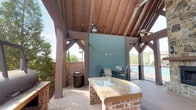 The Grilling Pavilion is a thoughtfully designed open-air structure built for outdoor entertaining and communal gatherings. The pavilion is anchored by a striking vaulted ceiling with exposed wood beams and tongue-and-groove planking, supported by sturdy timber-frame posts that lend the space a classic, rustic character. Ceiling fans are mounted overhead to provide comfortable airflow throughout the warmer months.

At the heart of the pavilion is a large barrel-style smoker grill, mounted on a custom brick pedestal, making it a focal point for grilling enthusiasts. Adjacent to the smoker is a built-in outdoor prep counter, also constructed with brick bases and a stone-topped surface, complete with a utility sink — ideal for food preparation and cleanup. The flooring throughout is a warm, classic brick paver surface that complements the overall aesthetic.

A seating area with metal chairs and a table is positioned near the back wall, offering a comfortable space to dine or relax. The pavilion overlooks a pool area, visible through a wrought iron fence, adding to the recreational atmosphere. Lush greenery and mature trees surround the perimeter, providing natural privacy and shade. The open-sided design ensures ample natural light and refreshing outdoor views from every angle.