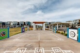 Located at 45760 W Village Pky in Maricopa, Arizona, this outdoor recreational amenity area is a well-designed community gathering space set within a modern residential development. The centerpiece of the space is a spacious concrete court marked with multiple game layouts, including shuffleboard lanes and hopscotch grids, making it a versatile destination for residents of all ages. A prominent pergola structure with white concrete columns and a warm wood-toned overhead lattice anchors the court, providing a shaded seating area with a bench positioned underneath. Colorful interactive signage panels in teal, green, and purple are installed along the court's perimeter, adding vibrant visual interest and community character to the space. Convenient waste receptacles are thoughtfully placed throughout the area. The court is bordered by open green lawn space that extends outward, offering an expansive, open-air feel under the wide Arizona sky. In the background, covered parking structures and the multi-story residential buildings of the surrounding community are visible, providing context for this amenity's role as a central outdoor hub. The overall ambiance is lively, community-oriented, and designed to encourage active outdoor engagement among residents.