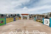 Located at 45760 W Village Pky in Maricopa, Arizona, this outdoor recreational amenity area is a well-designed community gathering space set within a modern residential development. The centerpiece of the space is a spacious concrete court marked with multiple game layouts, including shuffleboard lanes and hopscotch grids, making it a versatile destination for residents of all ages. A prominent pergola structure with white concrete columns and a warm wood-toned overhead lattice anchors the court, providing a shaded seating area with a bench positioned underneath. Colorful interactive signage panels in teal, green, and purple are installed along the court's perimeter, adding vibrant visual interest and community character to the space. Convenient waste receptacles are thoughtfully placed throughout the area. The court is bordered by open green lawn space that extends outward, offering an expansive, open-air feel under the wide Arizona sky. In the background, covered parking structures and the multi-story residential buildings of the surrounding community are visible, providing context for this amenity's role as a central outdoor hub. The overall ambiance is lively, community-oriented, and designed to encourage active outdoor engagement among residents.