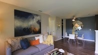 This single-floor space showcases a thoughtfully designed open-concept layout that connects the living and dining areas in a fluid, functional arrangement. The living area is anchored by a plush gray sofa adorned with an eclectic mix of decorative pillows in navy, orange, and neutral tones, complemented by a cozy yellow throw blanket. A striking large-scale abstract painting in deep blues and warm golds dominates the wall above the sofa, serving as a bold focal point that sets the artistic tone of the space. Rich, dark hardwood flooring runs throughout, adding warmth and visual continuity between areas. The dining space, visible from the living room, features a glass-top table paired with sleek white chairs and a geometric gold mirror that reflects light and adds a glamorous accent. A ceiling fan is positioned overhead, contributing to the room's comfort. Neutral gray walls provide a versatile backdrop that allows the colorful furnishings and artwork to stand out. The overall ambiance is contemporary and curated, balancing bold design choices with a welcoming, lived-in feel.