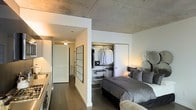 This single-floor living space showcases a refined industrial aesthetic, anchored by a striking exposed concrete ceiling that lends an urban, loft-like character to the interior. The sleeping area is thoughtfully integrated into the open-concept layout, featuring a generously sized bed dressed in neutral tones and flanked by soft ambient lighting. A built-in open wardrobe system provides practical, well-organized storage with dedicated hanging rails and shelving, neatly tucked behind a sliding panel for a clean visual finish. Dark hardwood flooring runs throughout, creating a warm contrast against the crisp white walls and raw concrete overhead. A framed shoji-style screen panel adds a subtle architectural detail, contributing to the space's layered design language. A corridor connects the sleeping area to additional spaces beyond, hinting at a well-considered floor plan. The overall ambiance is contemporary and sophisticated, blending modern minimalism with industrial textures to create a stylish and functional living environment.