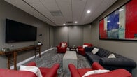 This well-appointed amenity space is thoughtfully divided into two distinct areas, each serving a unique purpose while maintaining a cohesive, contemporary aesthetic throughout. The lounge area features a sophisticated color palette of deep grays and bold red accents, with plush tufted seating including a charcoal Chesterfield sofa and vibrant red armchairs arranged around a concrete coffee table. Patterned carpet, recessed ceiling lighting, and a large-screen TV mounted above a console table create an inviting atmosphere for unwinding. A striking large-scale abstract artwork anchors the space, adding a dynamic visual focal point. The adjacent game room offers a bright, open contrast with light hardwood-style flooring, expansive floor-to-ceiling windows that flood the area with natural light and offer lush tree-lined views. A full-size ping pong table takes center stage, complemented by a foosball table and multiple high-top and standard dining tables for casual seating. Pendant lighting hangs above the game area, and tropical potted plants add a refreshing, natural touch. Together, these two areas form a well-balanced, multi-functional space ideal for both relaxation and active recreation.