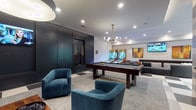 This well-appointed game room offers a diverse range of entertainment options across a single, thoughtfully designed floor. The main gaming area centers around a full-size pool table illuminated by a mid-century modern globe chandelier with brass accents, accompanied by a dedicated wall-mounted cue rack set against deep navy-painted paneling. Adjacent to the pool area, multiple skee-ball arcade machines and a shuffleboard table provide additional recreation, all overlooking a curved sectional sofa and wall-mounted flat-screen televisions ideal for sports viewing.

The flooring transitions between bold geometric checkered tile and soft area rugs, visually defining distinct zones throughout the space. Teal velvet accent chairs add a vibrant pop of color and serve as casual seating nodes. A private media nook features wood-paneled walls, built-in framed speakers, a large display screen, and plush sectional seating beneath bold artwork, creating an intimate screening environment.

A naturally lit dining area with floor-to-ceiling glass doors and a geometric tile floor provides a bright, casual gathering space. Throughout the room, curated artwork—including vinyl record wall installations and large-scale figurative paintings—reinforces the space's cultural, music-inspired character. Recessed lighting and pendant fixtures maintain a warm, welcoming ambiance throughout.
