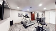 This fitness center is a thoughtfully designed workout space that provides a comprehensive range of exercise equipment for both cardio and strength training. The room features a neutral color palette with light grey walls and a patterned carpet flooring that adds a polished, professional feel to the environment. Recessed lighting throughout the ceiling ensures the space is well-lit, creating a bright and energizing atmosphere conducive to exercise. Two treadmills serve as the centerpiece of the cardio area, positioned side by side for easy access. Complementing the cardio section, a stationary bike is available for low-impact cycling workouts. A multi-function cable and weight machine is situated toward the far end of the room, offering a variety of strength training options. A warm-toned wood accent panel adds a touch of elegance to the space, balancing the functional equipment with refined design. Two framed black-and-white photographs adorn the walls, contributing a curated, sophisticated ambiance. The single-floor layout ensures everything is easily accessible, making this fitness center a convenient and well-appointed destination for a complete workout routine.