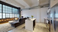 This single-floor urban loft masterfully blends industrial character with modern refinement. The open-plan living and kitchen area serves as the heart of the space, anchored by dramatic floor-to-ceiling factory-style windows framed in dark metal, set against exposed dark brick — a striking architectural feature that draws in abundant natural light. Soaring ceilings with exposed structural beams and visible HVAC ductwork reinforce the loft's industrial aesthetic, while polished concrete floors run throughout, lending a cohesive, contemporary feel.

The kitchen is well-appointed with warm wood-grain lower cabinetry, clean white upper cabinetry and open shelving, a subway tile backsplash, stainless steel appliances, and a sleek white island with bar seating — ideal for both cooking and casual dining.

A long hallway with wall sconces connects the living area to the private rooms. The bedroom is a calm, minimal retreat with soft neutral tones, track lighting, a large sliding-panel closet, and wood flooring. An in-unit laundry room with a full-size washer and dryer adds everyday convenience. The overall ambiance is sophisticated yet comfortable, balancing raw industrial elements with clean, modern finishes throughout.