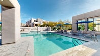 Located at 12100 N Mountain Centre Rd in Marana, Arizona, this outdoor amenity area at Legacy Apartments showcases a resort-style design that embraces the spirit of the Sonoran Desert. The centerpiece is a generously sized, crystal-clear swimming pool with sparkling turquoise water, complemented by an adjacent spa that invites relaxation year-round under the Arizona sun.

Surrounding the pool, a broad, tiled sundeck provides ample space for lounging, furnished with a collection of sleek chaise lounges and vibrant blue market umbrellas that offer both style and shade. A dedicated outdoor dining area with a table, chairs, and a large umbrella sits adjacent to the building's glass-fronted facade, ideal for al fresco gatherings.

The architecture reflects a modern Southwestern aesthetic, with smooth stucco exteriors, clean geometric lines, and flat-roof structures that echo the region's adobe building traditions. Desert-native landscaping, including towering saguaro cacti, desert shrubs, and mature trees, frames the space and enhances privacy. Green hedging along the perimeter further defines the space, while additional lounge seating tucked into quieter corners provides secluded spots for unwinding. The entire area is bathed in abundant natural sunlight, making it a vibrant and inviting outdoor retreat.
