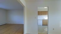 This single-floor residence is organized around a straightforward, functional layout that connects living and kitchen spaces through open archways and doorways. The living area features warm wood-tone flooring and white walls, creating a bright, neutral backdrop that allows for versatile furnishing arrangements. A rounded archway frames the transition between rooms, adding a subtle architectural detail to the otherwise clean-lined interior.

The kitchen is positioned adjacent to the main living space and is visible through a doorway, offering a sense of connectivity between areas. It features medium-toned wood cabinetry along the upper and lower walls, providing ample storage. A white dishwasher and a sink with a window above it are among the notable fixtures. The kitchen floor transitions to a lighter tile or vinyl surface, distinguishing it from the wood flooring in the living areas.

The white walls and ceilings throughout maintain a consistent, airy feel across the space. The overall ambiance is clean and understated, with a practical design that emphasizes usability and ease of movement between the main areas of the home.