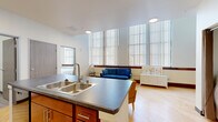 This thoughtfully designed 1-bedroom, 1-bathroom unit offers a functional and comfortable layout across a single floor. The open-concept kitchen and living area forms the heart of the space, where a central island with a double sink and dark countertop serves as both a prep station and casual gathering point. The kitchen is equipped with warm-toned wood cabinetry, black appliances including a range and full-size refrigerator, and a decorative tile backsplash. Light hardwood flooring flows seamlessly throughout, enhancing the bright and airy atmosphere. The living area benefits from an impressive bank of large windows fitted with vertical blinds, allowing abundant natural light while offering privacy control. High ceilings in the living space further amplify the sense of openness. The cozy bedroom features a window with outdoor tree views, continuing the warm hardwood flooring. The full bathroom is designed with accessibility in mind, incorporating grab bars, a shower with curtain, and easy-care luxury vinyl plank flooring. A dedicated closet and in-unit washer and dryer add practical convenience. The overall ambiance is clean, modern, and welcoming.