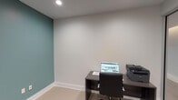This business center offers a well-appointed, single-floor workspace designed to support professional productivity in a clean and modern environment. The space is thoughtfully arranged with multiple individual workstations, each equipped with a desk, ergonomic office chair, and laptop computer. One workstation also features a multifunction printer, providing convenient document handling capabilities.

The interior design balances a neutral, professional palette with subtle pops of color. One workspace is anchored by a striking teal accent wall, adding a touch of vibrancy, while other areas feature crisp white and light gray walls that contribute to a bright, focused atmosphere. Recessed ceiling lighting throughout ensures consistent, even illumination across all workstations.

A standout architectural feature is the full-height glass partition wall with sleek aluminum framing, which separates the business center from an adjacent corridor. This transparent boundary allows natural light to flow into the space while maintaining a sense of openness and visibility. Light tile flooring with a wood-plank pattern runs throughout, complementing the contemporary aesthetic.

The space is accessed through a solid wood-tone door visible beyond the glass partition, and clean white baseboards frame the room neatly. Overall, the business center provides a functional, polished, and inviting environment suited for focused work tasks.