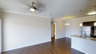 This single-floor space is designed with a clean, open layout that promotes a natural flow between the living, dining, and kitchen areas. The living area is finished with rich, dark hardwood flooring that adds warmth and depth to the space, while crisp white walls and crown molding lend an elegant, polished character throughout. A ceiling fan with a built-in light fixture keeps the living area comfortable year-round, complementing the well-distributed recessed lighting overhead. The dining area is distinguished by a classic chandelier, providing a refined focal point that defines the space without interrupting the open feel. Adjacent to the dining area, the kitchen features granite countertops, partially visible at the edge of the frame, hinting at quality finishes carried throughout the home. A hallway leading to additional rooms can be seen in the background, suggesting a thoughtfully designed floor plan with clear separation between living and private spaces. Neutral tones across the walls and trim create a versatile backdrop, making the space easy to personalize. Overall, the interior strikes a balance between functional design and understated elegance, making it a comfortable and visually cohesive single-level home.