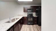 This two-floor residence is designed with a clean, modern aesthetic and a practical layout that makes efficient use of space throughout both levels. The main floor features wood-style plank flooring that extends through the living area and into the kitchen, creating a cohesive and easy-to-maintain foundation. A staircase connects the two floors, positioned alongside a hallway that leads to additional rooms and storage. The kitchen is well-equipped with rich dark cabinetry, granite-style countertops, a full suite of black appliances including a refrigerator, range, and built-in microwave, and a stainless steel sink. A peninsula counter separates the kitchen from the adjacent living space, adding both function and definition to the open layout. A half bathroom on the main level includes a dark-cabineted vanity with a granite countertop and wood-style flooring. Upstairs, carpeted bedrooms provide a comfortable, quieter retreat, with windows allowing in natural light. A walk-in closet with dual hanging rods offers generous storage. The full upper bathroom features a tub-shower combination, a dark vanity with granite countertop, and tile flooring. A dedicated laundry area with washer and dryer hookups rounds out the upper level.