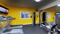This fitness room delivers a focused and energetic workout environment, immediately distinguished by its vivid yellow accent walls that create a motivating atmosphere. The space is finished with durable rubber flooring, providing both safety and comfort during exercise. A full-length mirror is mounted along one wall, allowing users to monitor their form while training and visually expanding the room. The centerpiece of the space is a multi-function cable machine, suitable for a wide range of strength training exercises. A workout bench is positioned nearby, complementing the resistance equipment for a well-rounded routine. A wall-mounted oscillating fan ensures adequate airflow and comfort during intense sessions. A single window along the yellow wall introduces natural light and offers a glimpse of the surrounding greenery outdoors. Overhead fluorescent lighting provides consistent illumination throughout. The textured ceiling and clean white baseboards add a neat, finished look to the space. Compact yet well-equipped, this fitness room provides the essential tools needed for an effective strength and conditioning workout.