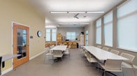 Located at 9393 Tidwell Rd in Houston, TX, this single-floor community facility is thoughtfully designed to serve a variety of educational and social functions. The space features an open, airy layout with warm yellow accent walls, neutral gray tones, and durable flooring throughout, creating a welcoming and functional environment.

The main activity area is generously proportioned, accommodating multiple white rectangular tables and modern stackable chairs arranged to support group gatherings, workshops, or collaborative sessions. Overhead linear lighting and ceiling fans ensure the area remains well-lit and comfortable. Large windows fitted with roller shades allow ample natural light to fill the room.

A dedicated kitchen area lines one wall and is equipped with stainless steel appliances including a full-size refrigerator, dishwasher, and microwave, complemented by light maple cabinetry and a continuous countertop with a sink — making it well-suited for community meals or events.

A children's corner provides a nurturing, age-appropriate space outfitted with small wooden tables and chairs, a bookshelf stocked with learning materials, and playful wall décor. A cozy seating area with a compact sofa offers a comfortable spot for reading or supervision.

A computer workstation area with desktop computers and ergonomic seating supports digital learning and productivity. Together, these distinct zones make this facility a comprehensive, community-centered resource.