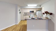 This single-floor residence offers a thoughtfully designed layout that balances comfort and functionality. Throughout the space, warm hardwood-style flooring creates a cohesive and inviting atmosphere, complemented by neutral-toned walls that allow for versatile styling.

The open-concept living area is spacious and bright, with natural light filtering in through a sizable window and a ceiling fan overhead providing year-round comfort. Tasteful artwork adorns the walls, adding character and visual interest to the space. A cozy nook features a wicker basket with plush pillows, making it an ideal reading or relaxation corner.

The kitchen is a standout feature, offering ample cabinetry with light wood finishes and generous granite-style countertops that extend into a peninsula breakfast bar — ideal for casual dining or meal prep. The kitchen is equipped with white appliances including a range and refrigerator, as well as a stainless steel microwave and sink, providing all the essentials for comfortable living.

The entryway is open and welcoming, with clean sightlines across the main living area. Multiple doorways lead to additional rooms, suggesting a practical and well-organized floor plan. The consistent flooring and soft color palette throughout the space contribute to an overall sense of calm, modern living.