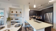 This single-floor residence showcases a contemporary design with a cohesive aesthetic carried throughout. The kitchen is outfitted with dark espresso cabinetry extending to the ceiling, a tile backsplash, stainless steel appliances including a refrigerator, electric range, and built-in microwave, plus light quartz countertops. A peninsula with an undermount sink and bar seating connects the kitchen to the dining area, where pendant lighting and open decorative wall shelving add architectural interest. Wood-look flooring flows through the main living areas, anchoring the open-concept layout. The living area provides a bright, comfortable retreat with clean white walls and ample room for seating. The bedroom is a standout space, featuring large windows framing scenic water and tree views, a ceiling fan, and wood-look flooring. The bathroom includes a dark-cabinetry vanity, large-format tile flooring, and a full soaking tub with shower combination. Directly accessible from the bathroom is a dedicated walk-in closet equipped with wire shelving and hanging rods, offering generous storage. The overall ambiance is modern and well-curated, with consistent finishes creating a polished, cohesive feel throughout.