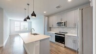 This thoughtfully designed one-bedroom, one-bathroom unit offers a efficient and modern living experience across a single floor. The open-concept layout seamlessly connects the kitchen and living area, creating a bright and cohesive space. The kitchen is a standout feature, showcasing gray shaker-style cabinetry, a herringbone tile backsplash, crisp white quartz countertops, and a peninsula with an undermount sink. A trio of matte black pendant lights adds a contemporary design touch overhead. Stainless steel appliances — including a refrigerator, range, microwave, and dishwasher — complete the well-appointed kitchen. The living area extends naturally from the kitchen, with natural light filtering in through glass-paneled doors and windows fitted with blinds. Warm hardwood-style flooring flows consistently throughout the entire unit, adding cohesion and warmth. The bedroom is a comfortable, light-filled retreat with large windows and a ceiling fan. The bathroom features a white quartz vanity with gray cabinetry, wood-look flooring, and a tiled shower, with an adjacent walk-in closet providing generous storage. Built-in shelving near the entryway offers additional organizational convenience, completing this well-designed and functional home.