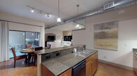 This well-appointed 2-bedroom, 2-bathroom residence offers a thoughtfully designed single-level floor plan that blends industrial-chic aesthetics with modern comforts. The open-concept kitchen is a standout feature, showcasing rich warm-toned cabinetry, granite countertops, black appliances, and an island with bar seating. Exposed ductwork and track lighting add an urban loft character to the kitchen and living areas. The living room is generously sized with hardwood flooring, an arched media niche, and large windows that allow natural light to fill the space. A dedicated dining area sits adjacent to sliding glass doors that open onto a private balcony, offering views of the surrounding brick exterior architecture. The primary bedroom features plush carpeting, a ceiling fan, and direct access to its en-suite bathroom, which includes a dual-sink vanity with granite countertops, a separate glass-enclosed shower, and a soaking tub. The second bedroom is equally well-sized, with carpet flooring, a ceiling fan, and ample natural light from a large window. The second full bathroom offers a tub-shower combination with tile surround. Polished concrete flooring accents select areas of the home, adding a modern industrial touch throughout this refined and functional residence.