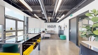 Located at 333 University Avenue in Kingston, Ontario, this single-floor space offers a compelling blend of industrial design and modern comfort. The open ceiling reveals exposed ductwork, conduit, and linear lighting fixtures painted in a dark finish, lending the space a contemporary, loft-like character. Polished concrete floors run throughout, providing a clean, durable foundation that complements the overall aesthetic.

The main open area is anchored by a bold yellow foosball table, setting an energetic and social tone. Adjacent to it, a comfortable lounge zone features a generous sectional sofa, ottoman, and coffee table arranged beneath a large flat-screen TV and custom wall mural branded with "Unity," adding a distinct artistic focal point. Floor-to-ceiling windows with roller blinds line the perimeter, flooding the space with abundant natural light and offering views of the surrounding Kingston skyline and rooftop terrace access.

The kitchen is well-appointed with grey cabinetry, white quartz countertops, a reflective subway tile backsplash, and stainless steel appliances including a range, dishwasher, and microwave. Ample counter space and storage make it fully functional for communal use. Together, these elements create a vibrant, multipurpose space ideal for collaboration, relaxation, and socializing.