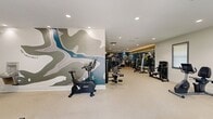 This 1,170-square-foot fitness center is a thoughtfully designed single-floor facility offering a comprehensive workout environment. The space is divided into distinct functional zones, making efficient use of its open layout. The cardio area features multiple treadmills, a rowing machine, stationary bikes, and an elliptical trainer, all positioned near large windows fitted with roller shades that allow ample natural light to fill the room. A set of glass doors provides direct outdoor access, further enhancing the bright, open atmosphere. The strength training zone is a standout feature, anchored by a full-wall mirror with elegant accent lighting above, reflecting a well-stocked dumbbell rack, a multi-station cable machine, a weight bench, and a leg press machine. An accent wall painted in sage green adds a modern, calming aesthetic, complemented by a decorative topographic mural. Recessed ceiling lighting throughout ensures consistent illumination across the entire space. The neutral-toned, durable flooring ties the room together and supports high-traffic use. The facility is well-ventilated with ceiling-mounted HVAC vents and speakers. Overall, the fitness center provides a clean, modern, and fully equipped environment suitable for a wide range of workout routines.