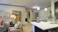 This well-appointed single-floor residence offers an open-concept design that thoughtfully integrates the living, dining, and kitchen areas into one cohesive and functional space. Light-toned hardwood-style flooring runs throughout, providing a warm and consistent foundation that ties the rooms together seamlessly.

The living area is generously proportioned, featuring neutral-toned walls that complement the warm ambient lighting. Decorative wall accents, including a distinctive scalloped arrangement, add artistic character to the space. A large ornate mirror with a natural wood frame enhances the sense of depth and brightness in the room.

The kitchen is designed with dark cabinetry contrasted by a clean white countertop peninsula, which doubles as a breakfast bar. Stainless steel appliances, including a full-sized refrigerator, add a modern, polished touch. Pendant lighting hangs above the peninsula, providing focused task lighting while contributing to the overall aesthetic.

The dining area is positioned centrally within the open layout, creating an effortless flow between cooking, dining, and relaxing. Corridor hallways lead to additional rooms, including what appears to be a bathroom with dark cabinetry visible in the background. Throughout the space, the warm lighting and carefully considered design details create an inviting and comfortable ambiance.